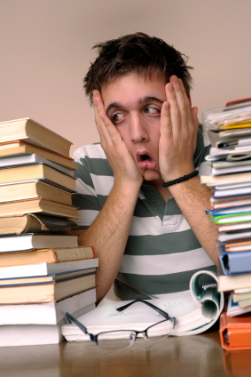 An overwhelmed student sits at a desk with his head in his hands, flanked by two towering stacks of textbooks