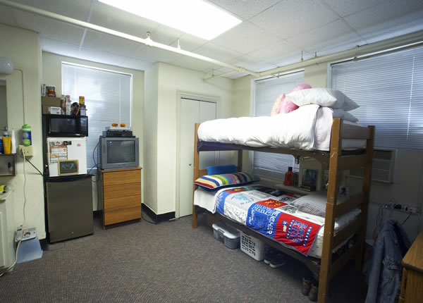A typical dorm room with a two-story bunk bed, an old tv, a fridge and a closet. The room is somewhat neat, with a few personal items belonging to a student.