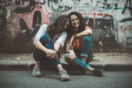 Two girls seated close together on a sidewalk, their faces lit up with cheerful smiles