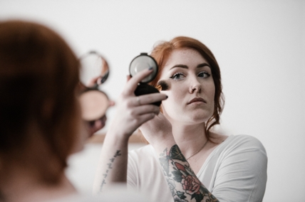 A woman holds a makeup applicator to her face and looks at her reflection in a mirror